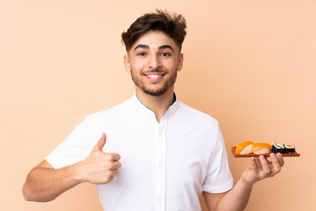 Homem árabe comendo sushi isolado em uma parede bege com o polegar para cima porque algo bom aconteceu