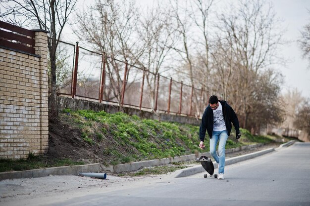 Homem árabe de estilo de rua em óculos com longboard longboard na estrada