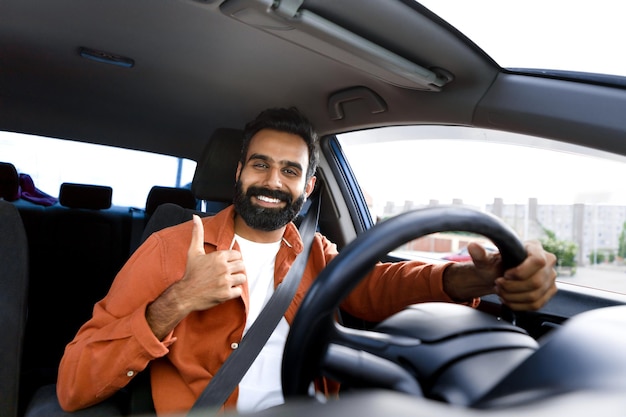 Homem árabe feliz dirigindo carro mostrando o polegar para cima sentado dentro