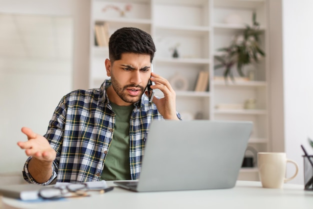 Homem árabe irritado falando no celular usando laptop