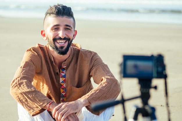 Homem árabe sentado na areia e falando em vídeo de tripé na praia conceito de criador de conteúdo de estilo de vida de cinegrafista de viagens