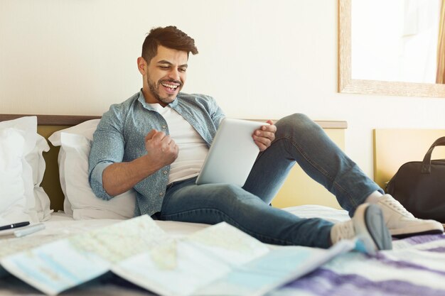 Homem árabe sentado na cama no hotel e usando o tablet digital. Cara empolgado reservando ingressos, planejando uma viagem de férias, copiando o espaço