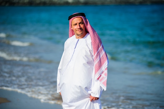 Homem árabe vestido de maneira do oriente médio posa em frente à costa do mar.