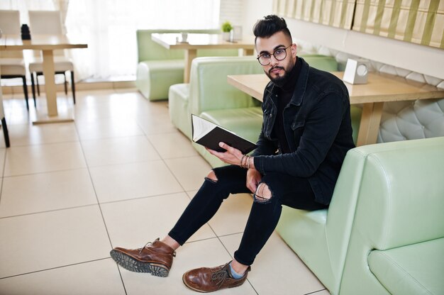 Homem árabe vestindo jeans preto e óculos sentado no café, lendo o livro