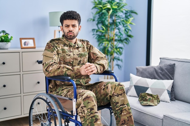 Homem árabe vestindo uniforme do exército de camuflagem sentado na cadeira de rodas bochechas inchadas com cara engraçada. boca inflada de ar, expressão louca.