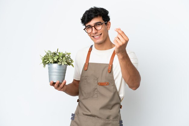 Homem argentino de jardineiro segurando uma planta isolada no fundo branco fazendo gesto de dinheiro