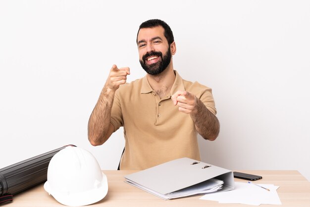 Homem arquiteto caucasiano com barba em uma mesa apontando para a frente com uma expressão feliz.