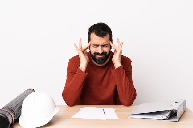 Homem arquiteto caucasiano com barba em uma mesa com dor de cabeça.