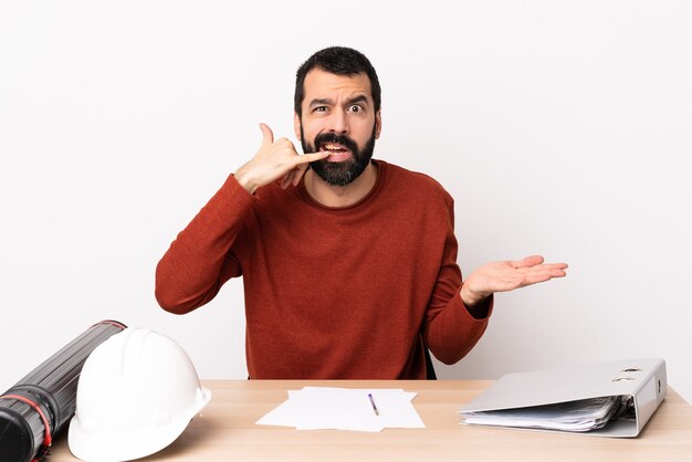 Homem arquiteto caucasiano com barba em uma mesa, fazendo gestos de telefone e duvidando.