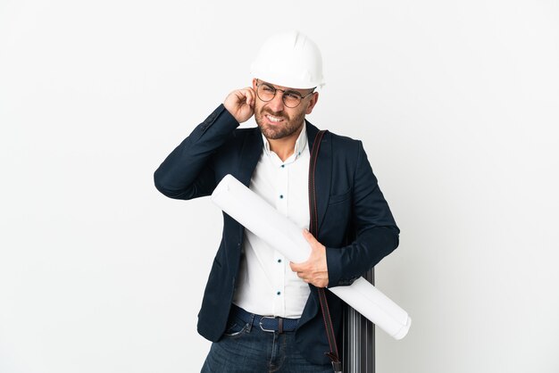 Homem arquiteto com capacete e segurando plantas isoladas no fundo branco frustrado e cobrindo as orelhas