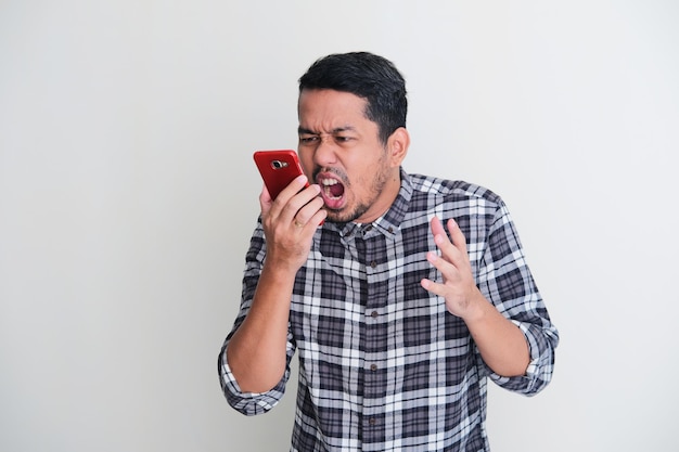 Homem asiático adulto gritando na frente de seu celular mostrando expressão de raiva