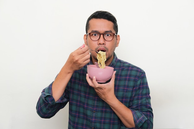 Homem asiático adulto mostrando expressão chocada ao comer uma tigela de macarrão