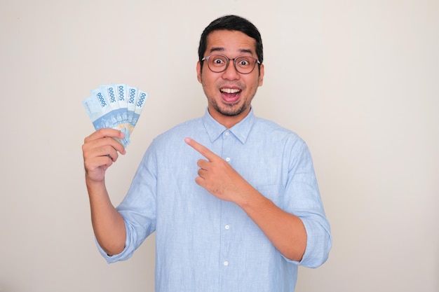 Homem asiático adulto mostrando expressão de rosto uau enquanto aponta para o dinheiro que ele segura