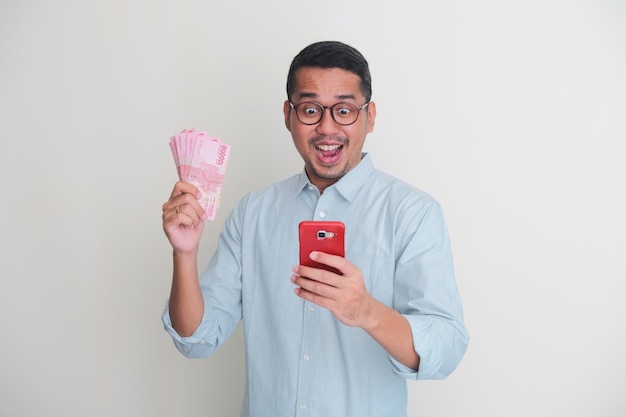 Homem asiático adulto mostrando expressão surpresa ao olhar para o celular enquanto segura papel-moeda
