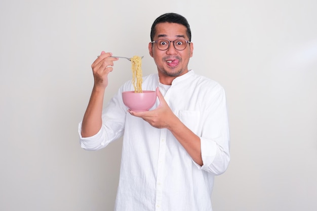 Homem asiático adulto mostrando uma expressão engraçada e animada enquanto come macarrão