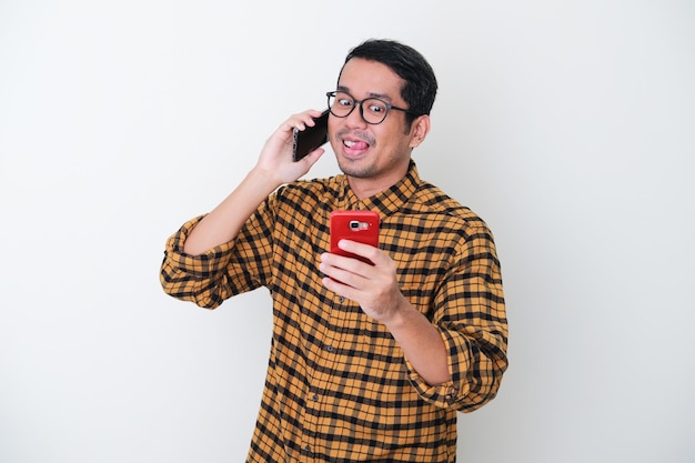 Homem asiático adulto mostrando uma expressão travessa ao ligar para alguém e enviar uma mensagem para outra pessoa ao mesmo tempo