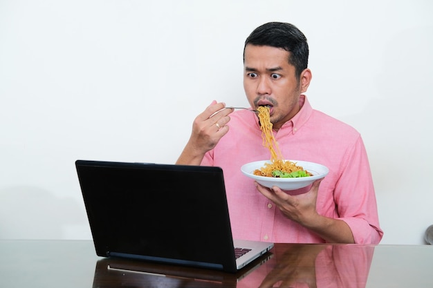 Homem asiático adulto olhando para seu laptop com expressão chocada enquanto come macarrão