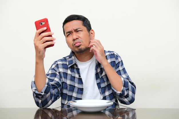 Homem asiático adulto sentado em frente ao prato vazio mostrando expressão confusa ao navegar no menu de comida em seu celular