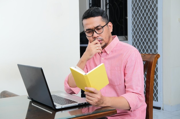 Foto homem asiático adulto sentado na frente de seu laptop enquanto lê um livro com expressão séria