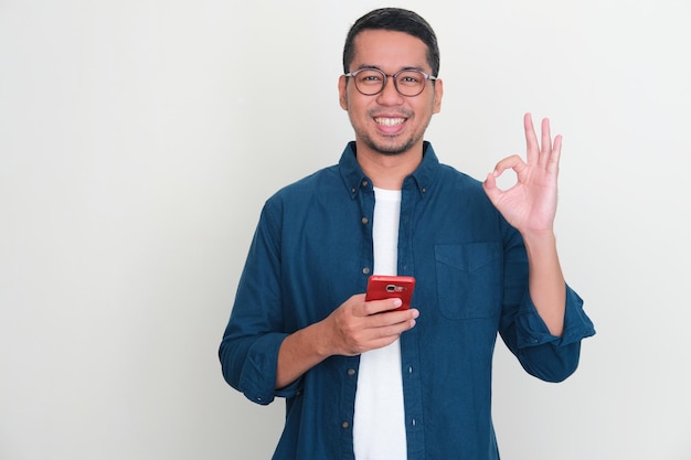 Homem asiático adulto sorrindo confiante e dando sinal de dedo OK enquanto segura um telefone celular