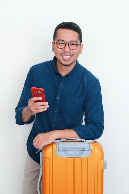 Homem asiático adulto sorrindo confiante segurando o celular posando na frente de sua bagagem