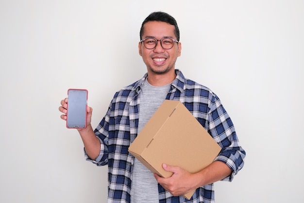 Homem asiático adulto sorrindo e mostrando a tela do celular em branco enquanto segurava um pacote de papelão