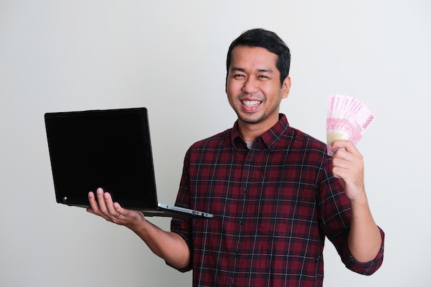 Foto homem asiático adulto sorrindo enquanto segura um laptop e dinheiro