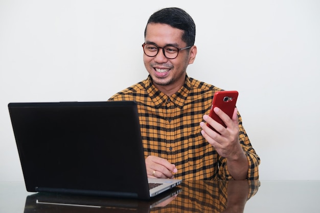 Homem asiático adulto sorrindo feliz ao olhar para seu laptop e segurando o celular
