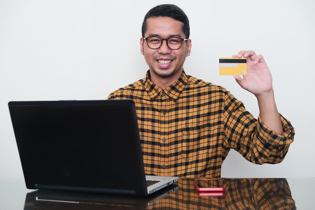 Homem asiático adulto sorrindo feliz e mostrando cartão de crédito enquanto está sentado na frente do laptop