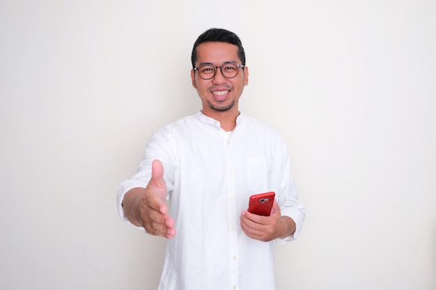 Homem asiático adulto sorrindo feliz e oferecendo um aperto de mão enquanto segura o celular