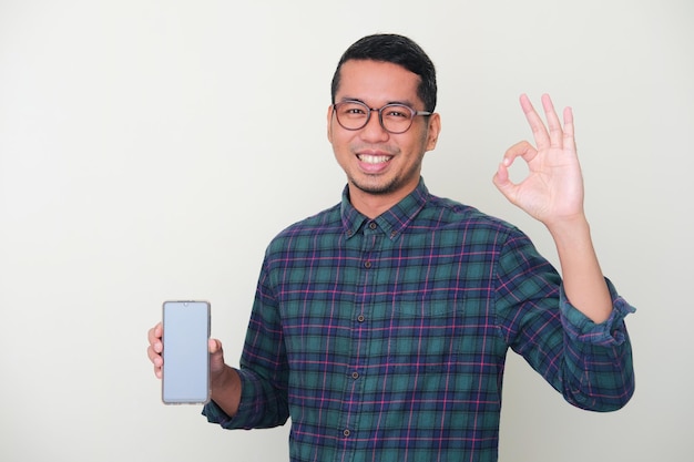 Homem asiático adulto sorrindo feliz enquanto mostra a tela do celular e dá sinal de OK