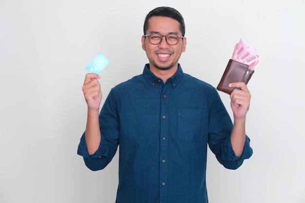 Homem asiático adulto sorrindo feliz enquanto segura o cartão de crédito em branco e a carteira cheia de dinheiro