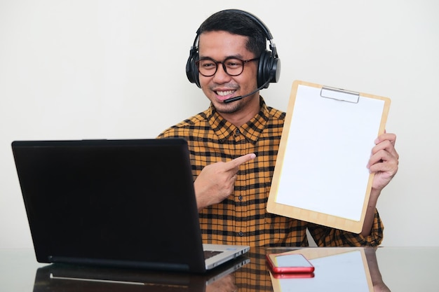 Homem asiático adulto usando fone de ouvido e mostrando papel branco vazio ao fazer reunião online usando laptop