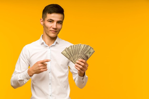 Homem asiático alegre em uma camisa branca aponta um dedo para dólares de dinheiro em uma imagem de fundo amarelo