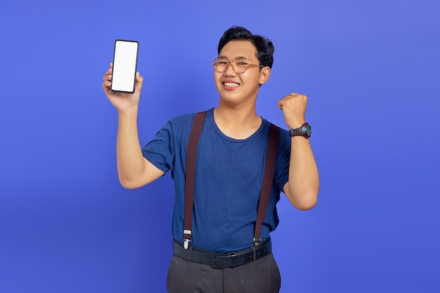 Homem asiático alegre mostrando a tela em branco do smartphone e celebrando a sorte sobre o fundo roxo