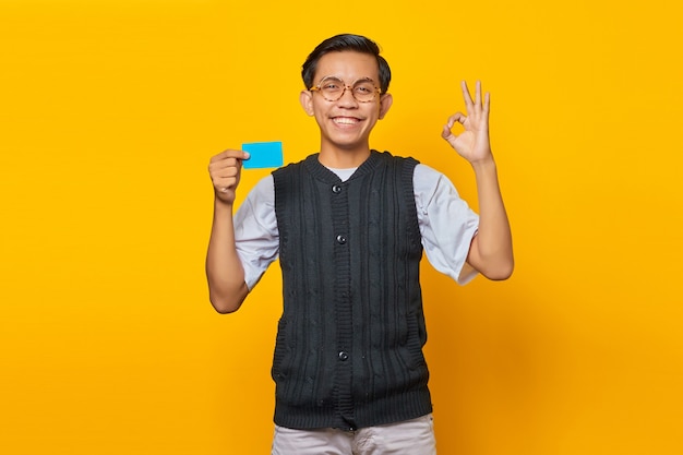 Homem asiático alegre mostrando o cartão de crédito e gesticulando sinal de tudo bem sobre fundo amarelo