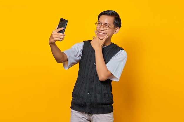 Homem asiático alegre usando telefone celular fazendo selfie sobre fundo amarelo