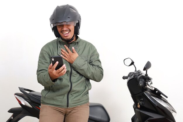 Foto homem asiático aliviado recebendo mensagem de seu telefone enquanto tocava seu peito perto de motocicleta isolado em branco