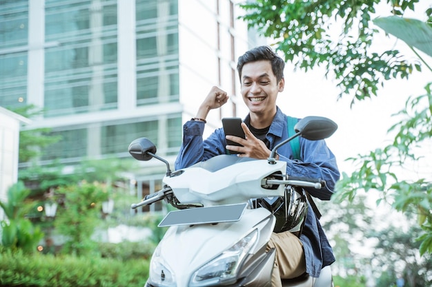 Homem asiático animado usando um celular enquanto andava de moto com edifícios no fundo da cidade