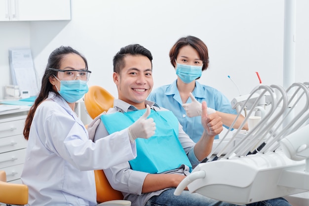 Homem asiático bonito e sorridente feliz, seu dentista e assistente com máscaras médicas mostrando o polegar para cima após terminar o tratamento