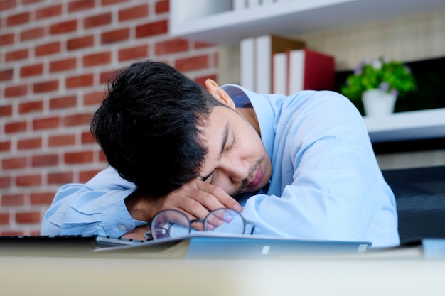 Homem asiático cansado dormindo na mesa de escritório. Jovem empresário com óculos sobrecarregados e adormeceu