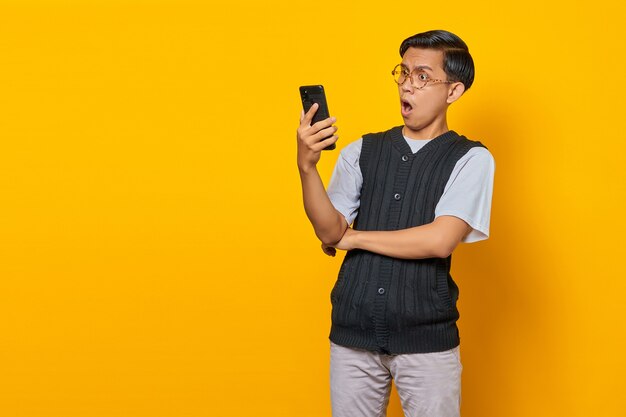 Homem asiático chocado olhando para um smartphone com a boca aberta sobre um fundo amarelo