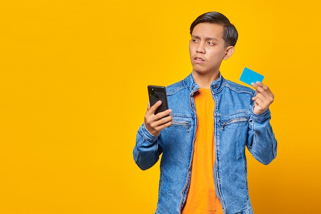 Homem asiático chocado segurando um telefone celular e mostrando um cartão de crédito sobre fundo amarelo