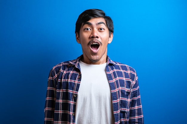 Homem asiático chocado vestido de casual ith boca aberta e mãos para cima olha para a câmera. jovem estudante asiático surpreso no fundo azul do estúdio