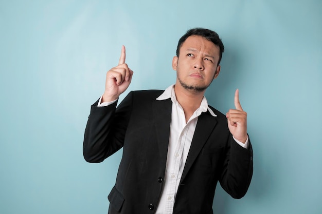 Homem asiático chocado vestindo terno apontando para o espaço de cópia acima dele isolado por fundo azul