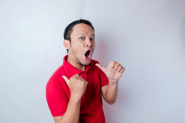 Homem asiático chocado vestindo uma camiseta vermelha apontando para o espaço de cópia ao lado dele isolado por um fundo branco