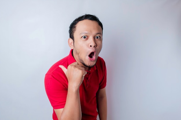 Homem asiático chocado vestindo uma camiseta vermelha apontando para o espaço de cópia ao lado dele isolado por um fundo branco