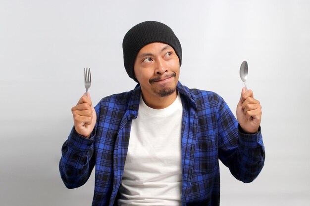 Homem asiático com fome morde os lábios enquanto segura um garfo e uma colher