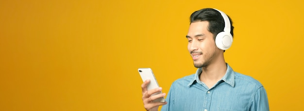 Homem asiático com fones de ouvido, segurando um telefone celular, sorrindo enquanto ouve música isolada em um fundo amarelo com espaço de cópia