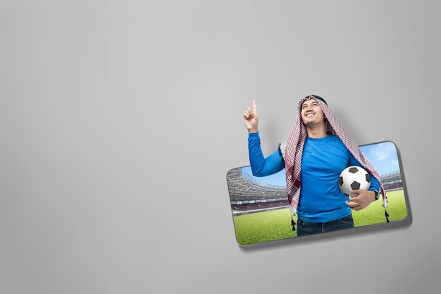 Homem asiático com keffiyeh em pé enquanto segura a bola com uma expressão animada no estádio de futebol